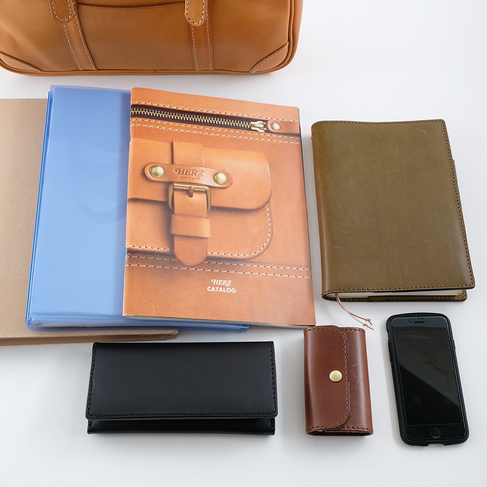 職人手作皮革肩背手提兩用公事包 BW-57 M，展示於此圖片中，搭配一本淺藍色文件夾、一本棕色皮革記事本、一本咖啡色皮革目錄、一個黑色長夾、一個咖啡色零錢包及一支黑色手機。