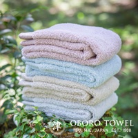 堆疊的毛巾，由左至右顏色為淺粉色、淺藍色、米白色和淺灰色。毛巾質地蓬鬆，邊緣有條紋裝飾。背景為綠葉模糊的自然環境。