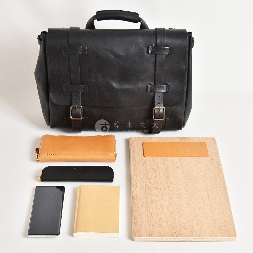 黑色皮革復古郵差包，附有黃褐色皮夾、黑色筆袋、手機、淺黃色筆記本和木質夾板。