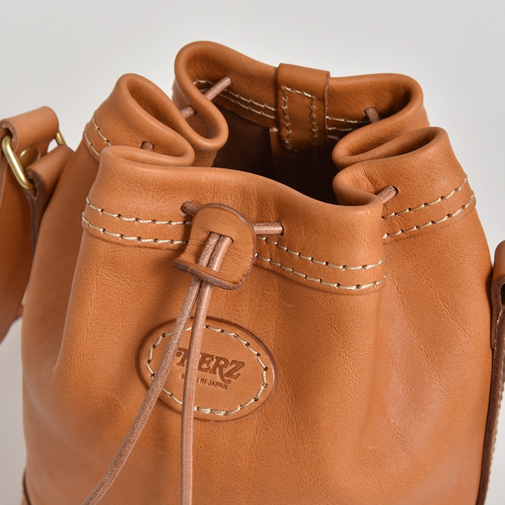 職人手作皮革束口單肩包，採用棕褐色皮革，配有米白色縫線和皮革束繩。包身有品牌壓印標誌，整體風格簡約。