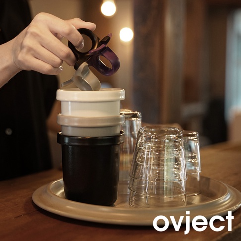 疊放的黑色、淺灰色和白色琺瑯馬克杯，杯口朝上，手中握持著一個紫色和銀色的握把，旁邊堆疊著透明玻璃杯，均放在金屬托盤上。