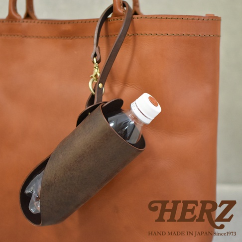 職人手作深棕色皮革Organ水壺套，附有長掛繩和金屬扣環，可掛於同色系皮革托特包上，中間裝有一瓶透明塑膠水瓶。