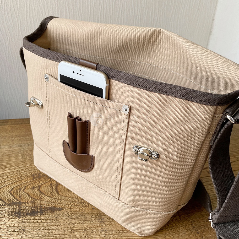 米色純綿帆布郵差包，配有棕色帆布包邊與肩帶。包身設有外置口袋，可放置手機，並有雙扣設計。