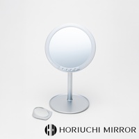 銀色金屬材質的桌上型化妝鏡，鏡面為圓形，鏡框下方有數個圓形觸控按鈕。鏡子底部為圓形底座，帶有支撐桿。一旁有一個小型的三角形放大鏡。