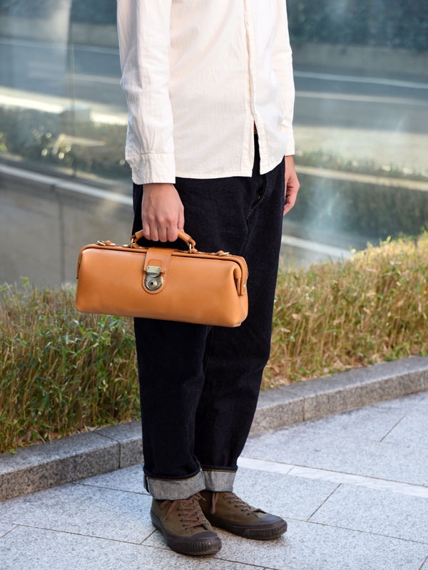 一只手拿著棕色皮革製的橫長形單肩包，包身結構硬挺，配有金屬鎖扣和提把，整體風格復古。