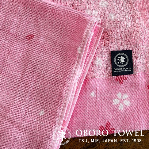 粉紅色紗布毛巾，帶有日式花朵圖案和 OBORO TSU, MIE, JAPAN EST. 1908 的品牌標籤