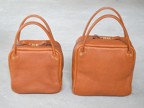 兩個棕色皮革方形托特包，一個較小，一個較大。兩者均有頂部提把、拉鍊開合和金色拉鍊頭。包包側面有金屬螺絲裝飾。