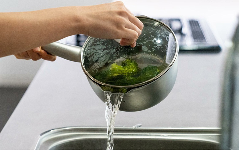 左手持用銀色不鏽鋼雪平鍋，鍋內有綠色花椰菜，鍋蓋為透明玻璃，正將鍋中水瀝入水槽。