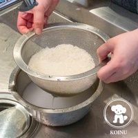 雙手正持著一個不鏽鋼濾水網，內部裝有米飯，濾網置於一個帶有刻度的不鏽鋼料理盆上方，水正從濾網中流出。