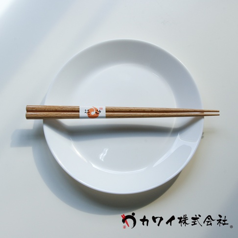 一雙天然原色木筷，末端有細緻的紋理，中間繫有印有橘色圖案和日文的白色紙帶，靜置於白色圓盤上。