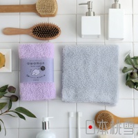 一條紫色、帶有品牌標籤的綿柔沐浴巾，旁邊是一條淺灰色、質地蓬鬆的沐浴巾。兩者皆為長方形，擺放在白色瓷磚背景上，周圍還有木柄刷、皂液瓶、牙刷和植物枝葉。