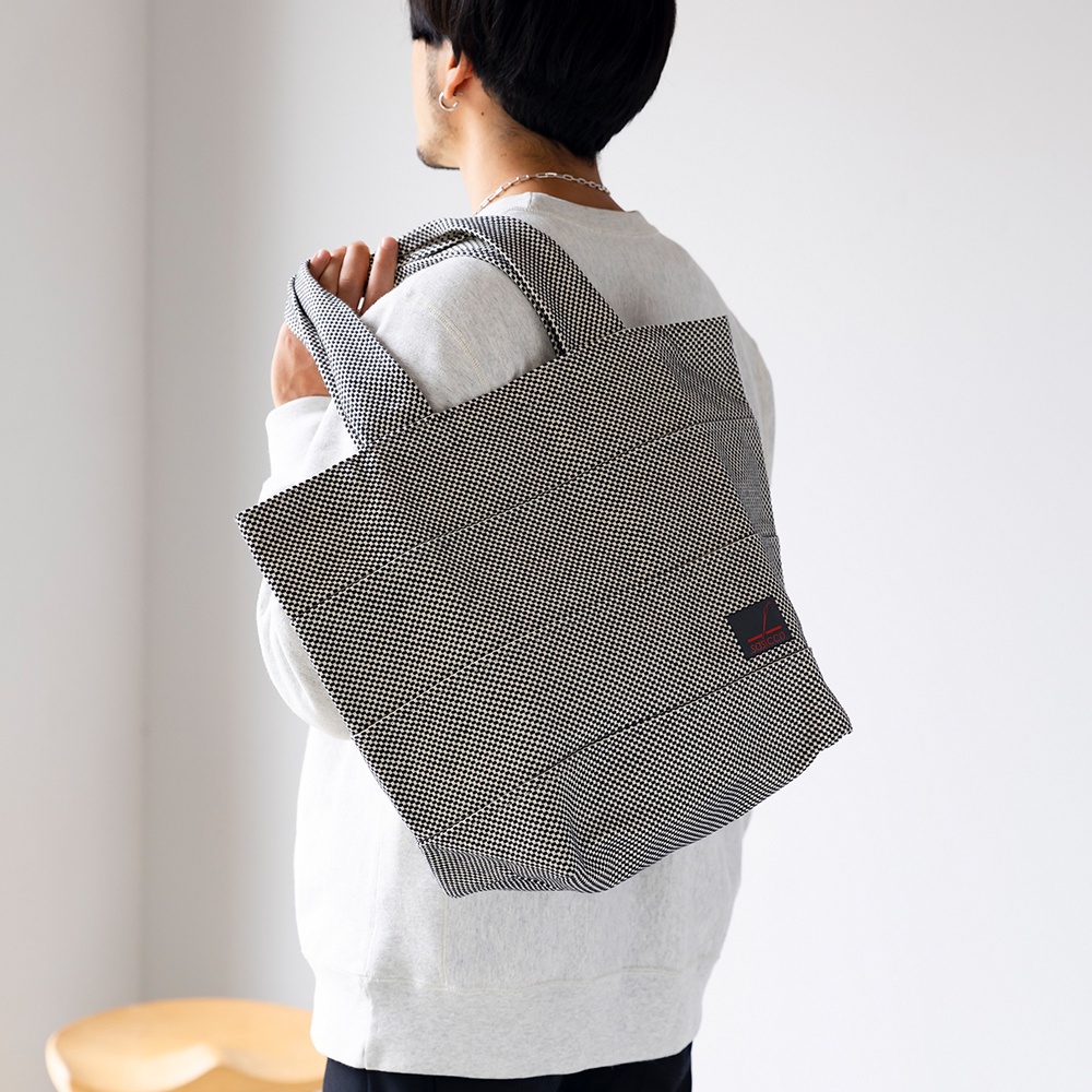 男士身穿淺灰色連帽衫，肩背一個黑色與白色細小格紋圖案的刺子織大容量托特包，包包右下角有一個紅色標籤。
