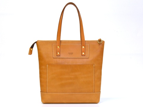 職人手作皮革威靈頓直式拉鍊托特包，採用天然棕色皮革製作，包身有垂直拉鍊開口，正面設有方形貼袋，提把處以鉚釘固定，整體風格簡潔經典。