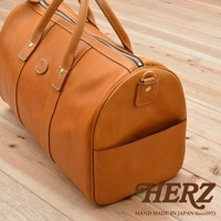 職人手作的棕色皮革波士頓包，具有圓筒形輪廓、頂部拉鍊開口、雙手提把、側邊插袋，以及金屬D型環肩帶連接點，搭配白色縫線。