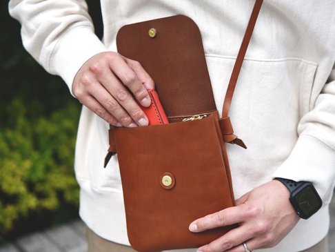 職人手作皮革橫式隨行袋，採棕色皮革材質，方形翻蓋設計，附有金色金屬釦。袋身簡約，可斜背，展示放入紅色卡夾的動作。