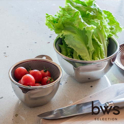 桌面上放置著一組 bws SELECTION 不鏽鋼備料濾網盆，其中較小的濾網盆裝滿新鮮紅番茄，較大的濾網盆內裝著洗淨的生菜。旁邊擺放著一把金屬廚房刀具與木質砧板，展現簡約實用的廚房備料場景。