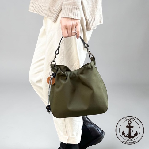 一位穿著淺色針織衫與白色休閒褲的模特兒，手提一個墨綠色防潑水材質的BALON mini手提包，包身呈現自然垂墜感，側面配有精緻的吊飾與繩扣設計，右下角帶有神戶clueto的錨型品牌標誌。