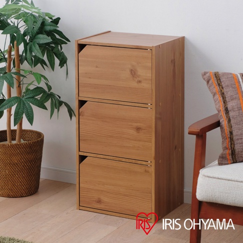 淺木色三層櫃，採用天然木紋材質，設有三個帶斜角門板的儲物空間，擺放在室內角落，旁邊為植物與椅子。