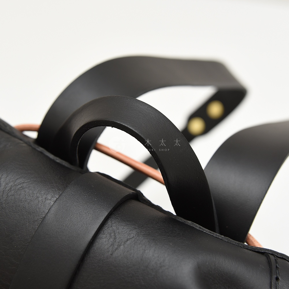 黑色皮革束口後背包特寫，可見圓潤的束口帶、皮革提把，以及細節處的銅色卯釘。