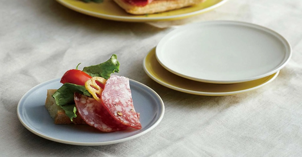 淺藍色美濃燒配菜盤上放著香腸、生菜和番茄，盤子邊緣略微上翹。背景有疊放的白色和黃色盤子。