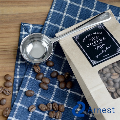 銀色金屬咖啡兩用量匙夾，配有一袋咖啡豆和藍色格紋布。