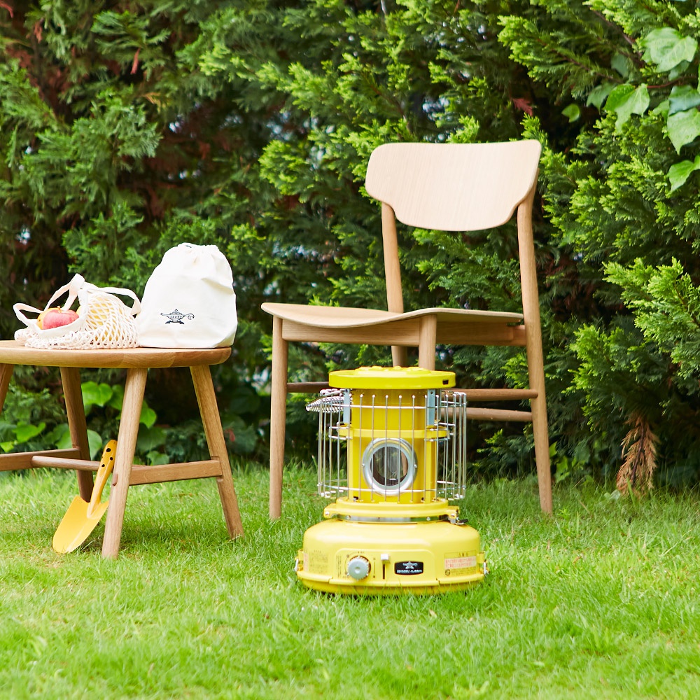 黃色卡式瓦斯暖爐，帶有金屬保護網和操作旋鈕，放置於草地上，背景為綠色樹叢和木製椅子。