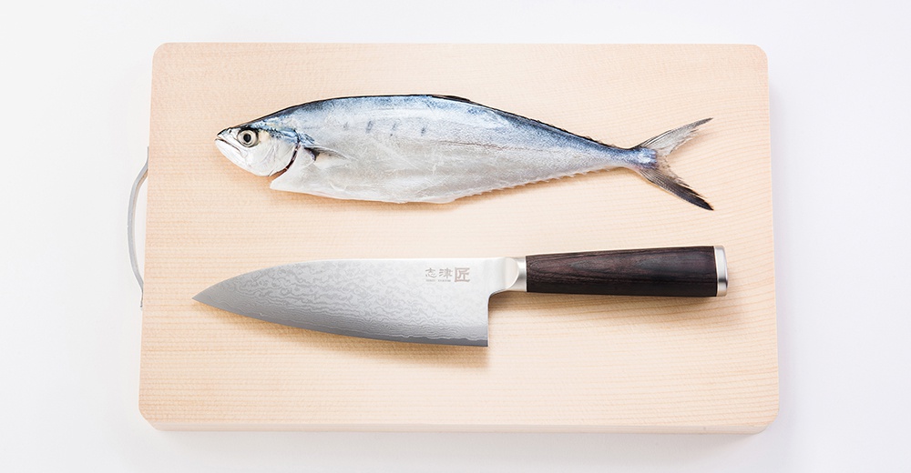 淺色木質砧板上，一條銀色帶有藍色斑紋的魚橫躺著，下方是一把 Santoku 萬用刀，刀身為大馬士革紋路，刀柄為深棕色木質，配有銀色金屬刀頸。