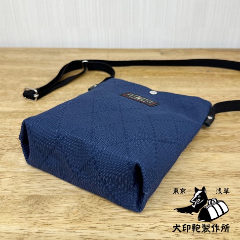 深藍色刺子織紋理的平板收納側背包，配有黑色可調節背帶，包身設有品牌標籤和金屬釦。