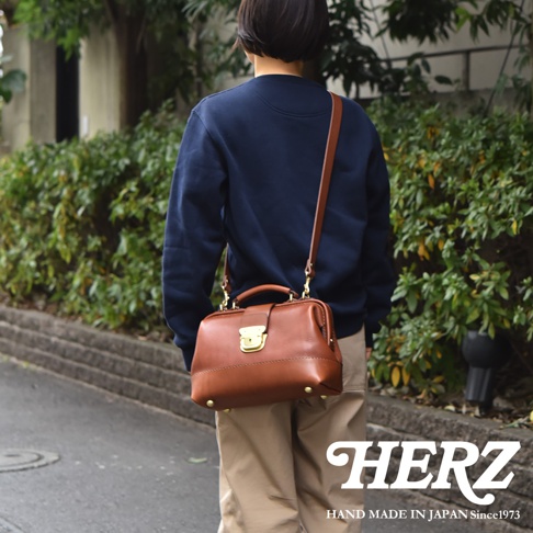 一位短髮女性背著日本HERZ品牌的棕色職人手作皮革兩用迷你口金包，包身正面裝有金屬扣環與手提把，搭配深藍色上衣與淺棕色長褲，於戶外綠色植栽旁行走呈現後背視角。