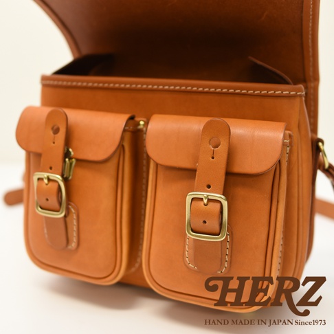 日本HERZ職人手作皮革經典雙扣郵差包，棕褐色全皮革材質，設有兩個翻蓋前袋，每個前袋皆有銅釦和皮帶設計，整體呈現復古手工質感。