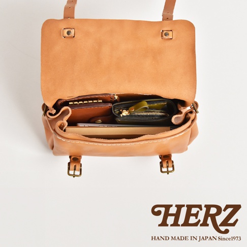 一個淺棕色皮革製作的復古郵差包，包包內裝有多個皮夾和卡夾，包包上方蓋子敞開，露出內部細節，包包正面有兩個金屬扣環。