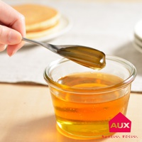 一只手拿著一個銀色金屬蜂蜜湯匙，湯匙的凹槽裡裝滿了濃稠的琥珀色蜂蜜。湯匙正從一個透明的玻璃罐中舀取蜂蜜，罐子裡也裝滿了蜂蜜。背景模糊，可以看到一些鬆餅。