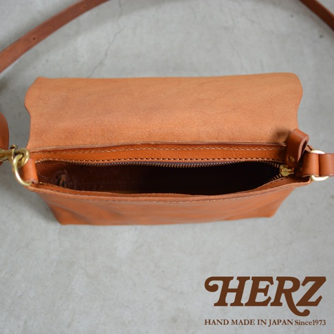 HERZ 品牌標誌，下方印有 HAND MADE IN JAPAN Since1973 字樣，圖片中央為一個棕色皮革斜背包，包蓋微開，露出拉鍊開口的內部。