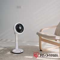 白色直立式循環扇，圓形扇頭，黑色扇網，可調式高度，放置於米色地毯上，旁邊有一張淺木色椅架、米色布面座椅的椅子。