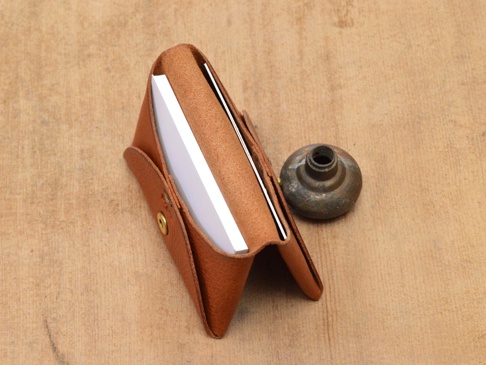 職人手作的雙開式卡片收納夾，採用棕色皮革材質，設有金色按扣，內部可收納白色卡片，旁邊擺放一個復古金屬旋鈕。