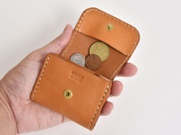 一只手拿著一個淺棕色皮革雙折收納零錢包，包內裝有數枚硬幣。零錢包有縫線細節和金屬按扣，表面印有「HERZ MADE IN JAPAN」字樣。