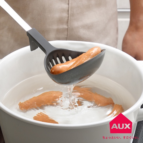 AUX 濾水湯匙正在從滾水中撈出兩根香腸，湯匙上有圓孔設計，白色鍋中有幾根香腸，右下角有 AUX 標誌和日文字樣。