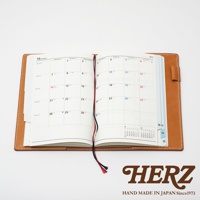 職人手作皮革B6記事本書套，展現天然皮革的溫潤質地，焦糖棕色皮革外殼，內含一本翻開的月曆筆記本，書本裝有紅藍色書籤帶，簡約實用。