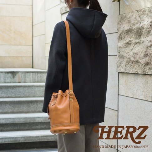 一位女士身穿深灰色連帽外套，肩背一個 tan 色皮革束口單肩包，包身有明顯的縫線和金屬鉚釘裝飾。