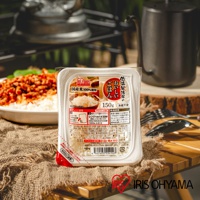 RIS FOODS 品牌標誌的低溫製法微波白飯，包裝上印有日文說明，標示「国産米100%使用」、「150g」、「低溫製法」等資訊。產品包裝為白色塑膠盒，部分透明。背景為戶外野營場景，有餐盤、餐具和炊具。