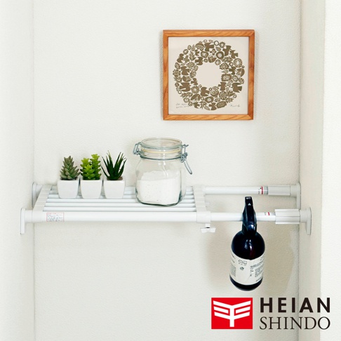 白色伸縮桿層架，架上擺放三個白色花盆內栽種的仿真植物、一個玻璃罐裝白色粉末，以及一個黑色噴霧瓶。層架下方懸掛著黑色噴霧瓶，上方牆面掛有一幅木框裝飾畫。