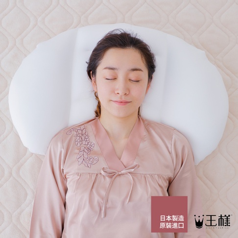一名女性仰躺在白色舒肩枕上，枕頭為雲朵造型，女性身穿粉色絲質睡衣，睡衣領口有花卉刺繡，背景為米色菱格紋床墊。