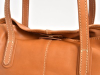 職人手作皮革橫式肩背托特包，採用溫暖的棕褐色皮革，搭配奶白色的縫線，展現細緻的工藝。包包的皮革表面光滑，部分細節處可見自然的紋理。