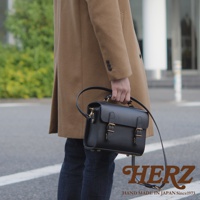 黑色皮革迷你兩用包，配有頂部提把和可拆卸肩帶，採用雙搭扣閉合設計，呈現職人手作的極簡風格。