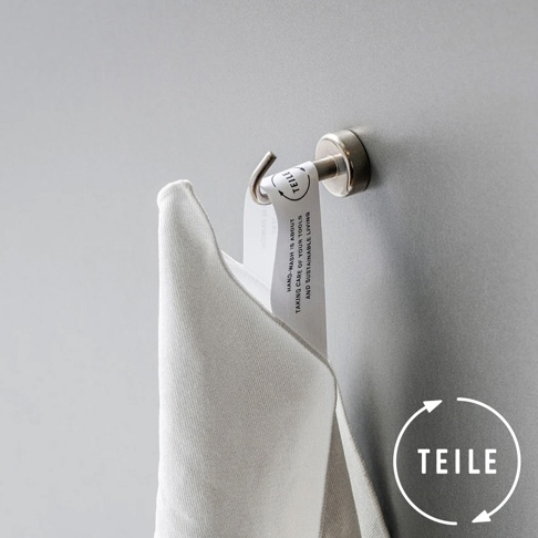 淺灰色纖維擦拭巾，質地柔軟，掛於金屬掛鉤上，標籤上印有黑色文字「TEILE」及英文說明，背景為淺灰色牆面。