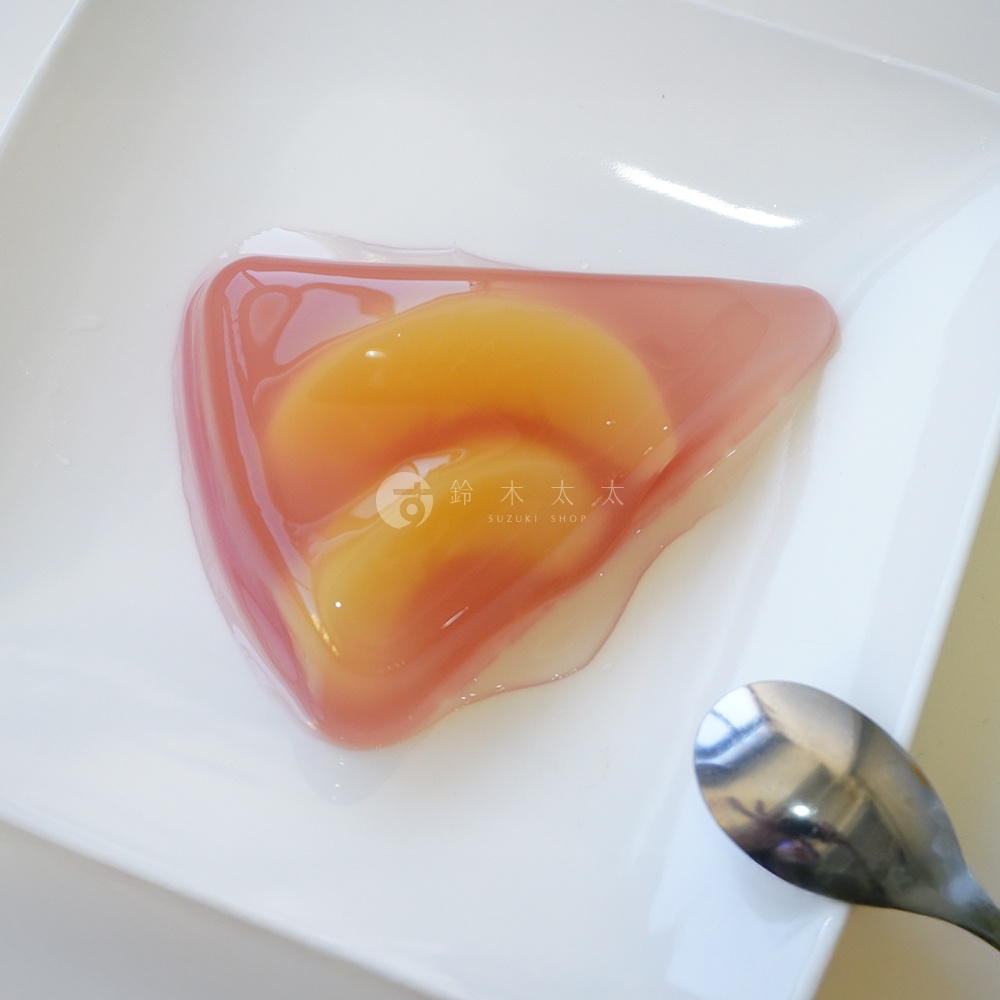 三角形粉紅色果凍中，包裹著兩片金黃色的水蜜桃果肉，果凍外層有水滴狀的透明凝膠，擺放在白色方形盤上，旁邊有一支金屬湯匙。