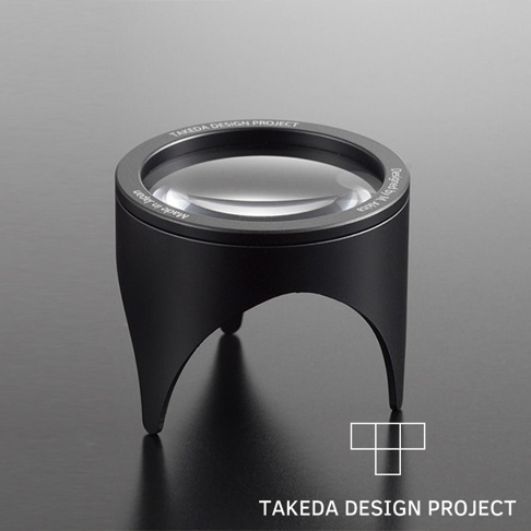黑色鋁製閱讀放大鏡，鏡片清晰，鏡框周圍刻有「TAKEDA DESIGN PROJECT」及「Made in Japan」字樣，底部有三個彎曲的支腳。