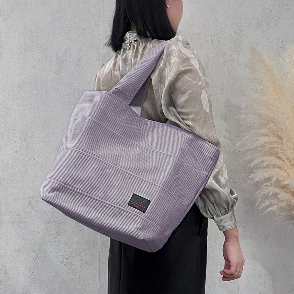 淺紫色三河木綿刺子織大容量托特包，帶有細緻織紋和黑色品牌標籤，展現簡約風格。