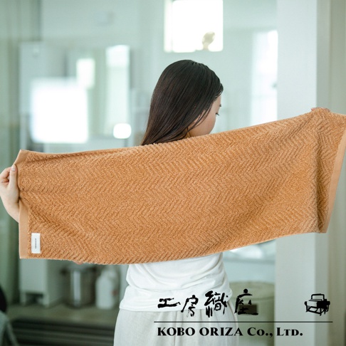一位女性展示一條橙棕色、人字紋織法的有機棉毛巾，毛巾質地柔軟，質感細膩。