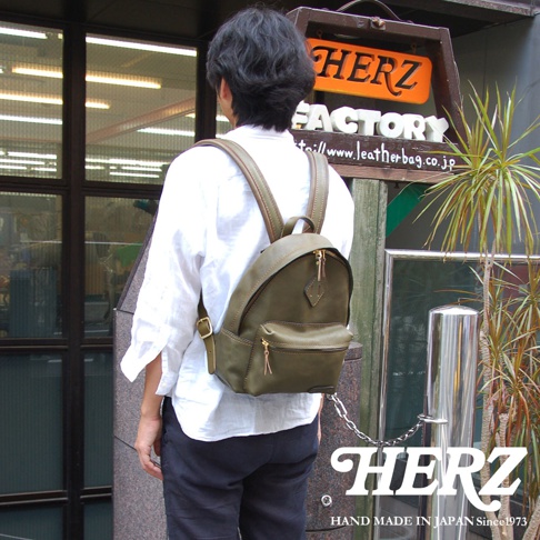 一位男士背著一個橄欖綠色的皮革後背包，背包上有金屬拉鍊和一個小前袋。